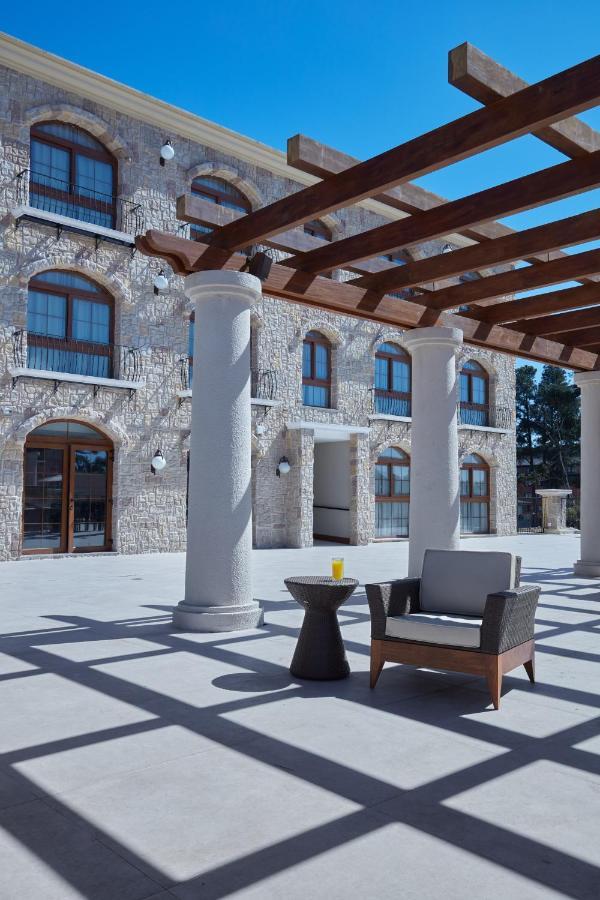 Buona Vitta Gramado Resort & Spa By Gramado Parks Zewnętrze zdjęcie The photo shows a well-designed outdoor space with a stone wall building in the background. There are several tall columns that support a wooden pergola overhead, creating a shaded area. In this space, there is a stylish chair and a small table besid