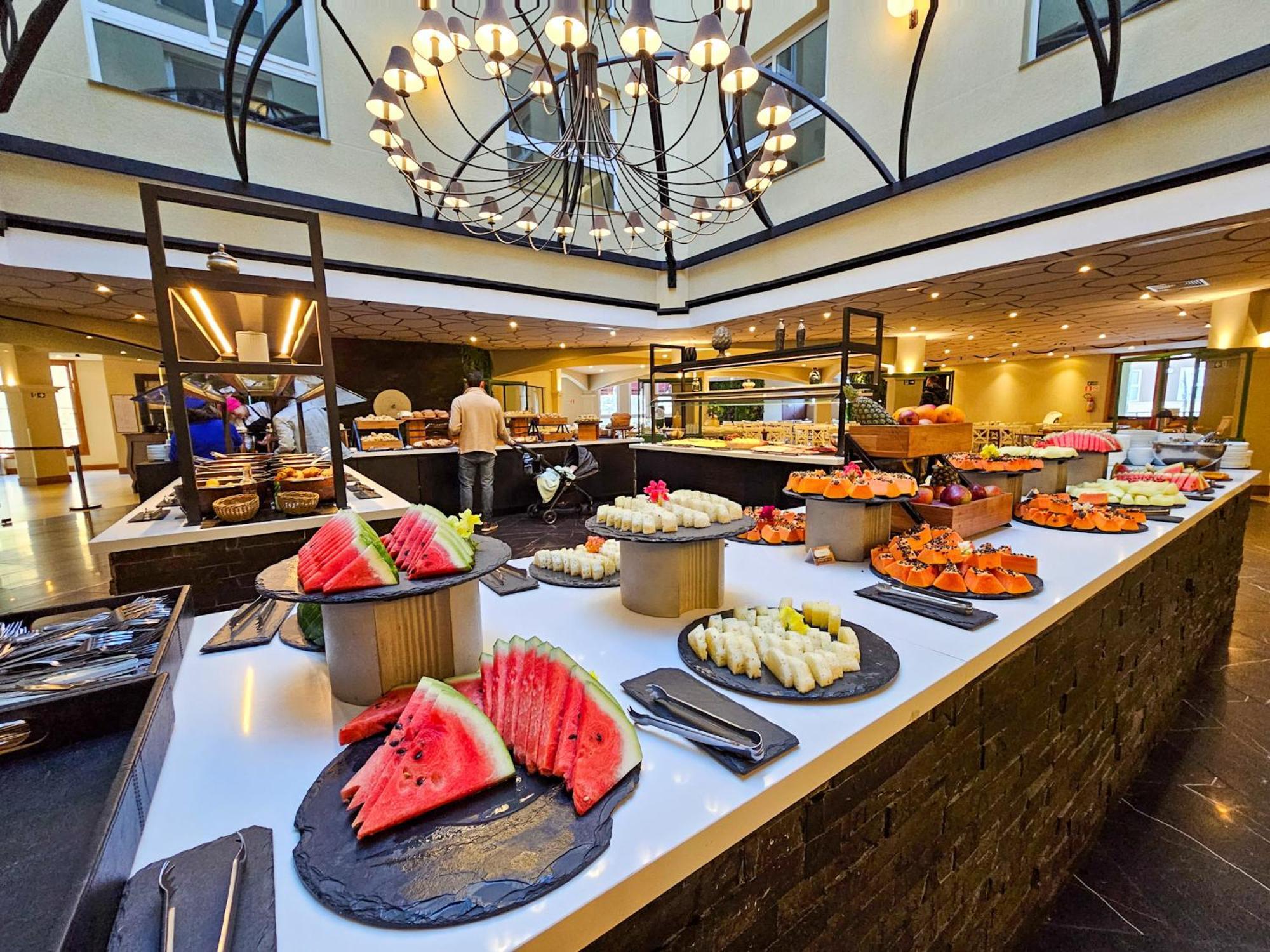 Buona Vitta Gramado Resort & Spa By Gramado Parks Zewnętrze zdjęcie The photo depicts a lavish buffet setup in a dining area. In the foreground, there are several platters featuring an assortment of colorful food items, primarily fruits like watermelon and various types of sushi or rolls. The buffet table is elegantl