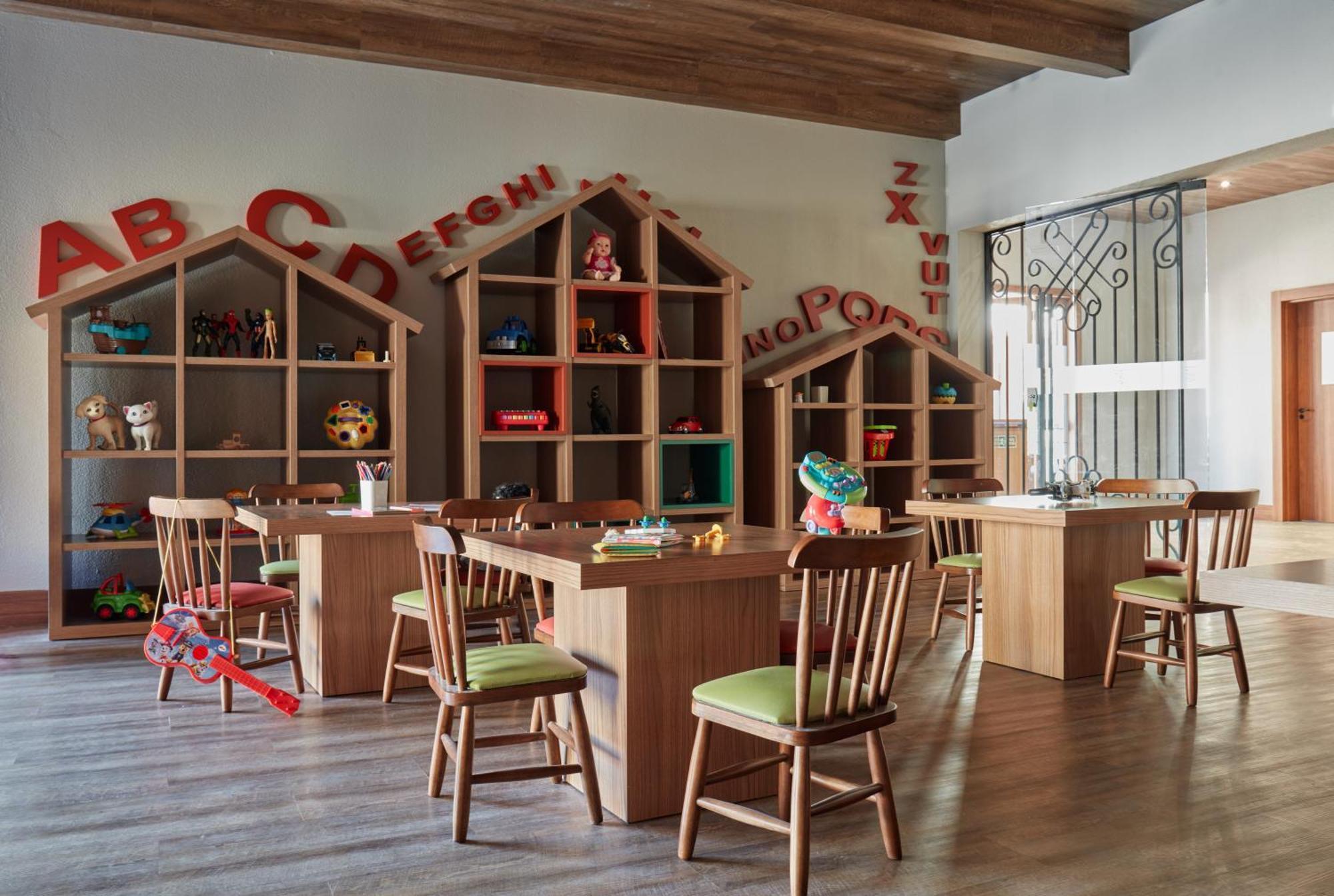 Buona Vitta Gramado Resort & Spa By Gramado Parks Zewnętrze zdjęcie The photo shows a classroom or playroom designed for children. It features wooden furniture, including several tables and chairs with green cushions. In the background, there are large, open shelving units shaped like houses, displaying various toys 