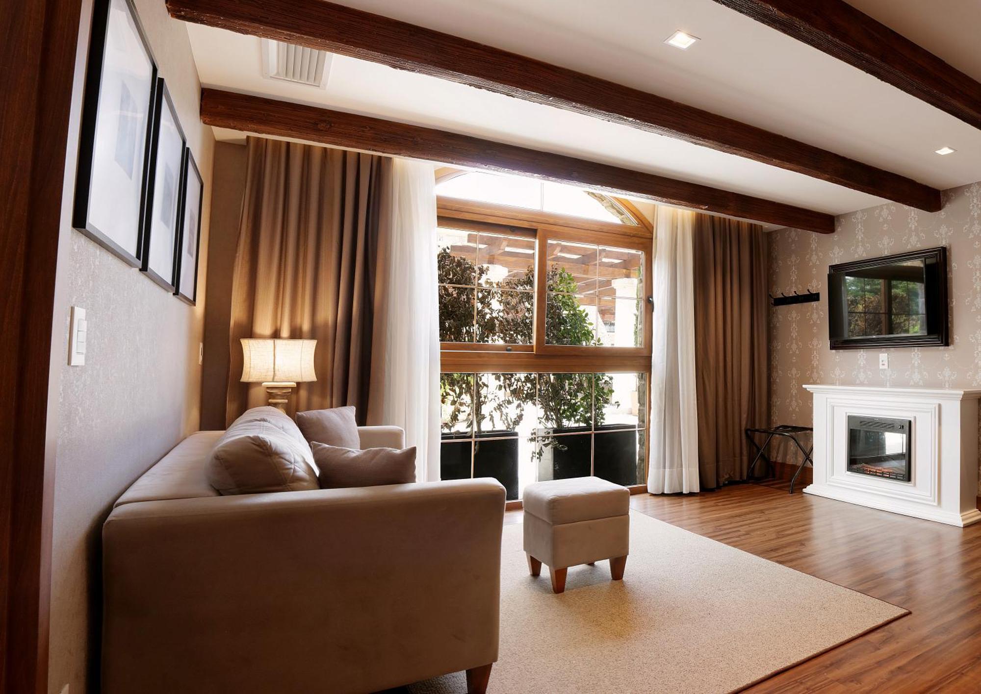 Buona Vitta Gramado Resort & Spa By Gramado Parks Zewnętrze zdjęcie The photo depicts a cozy living room setting. It features a light-colored couch, a small ottoman, and a floor lamp beside the couch. Large windows are covered with curtains, allowing natural light to illuminate the space. In the background, there is 
