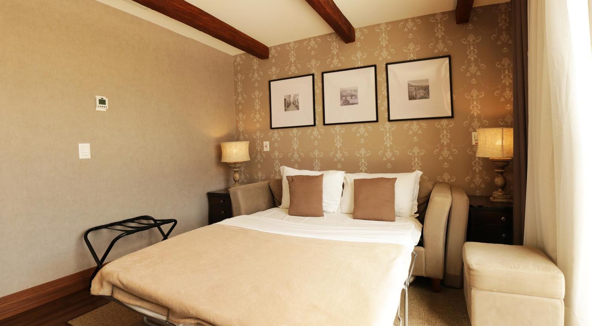 Buona Vitta Gramado Resort & Spa By Gramado Parks Zewnętrze zdjęcie The photo shows a cozy bedroom with a neutral color palette. There is a bed with white linens and two brown decorative pillows. On either side of the bed, there are bedside tables with lamps. The walls feature a subtle floral pattern, and three frame