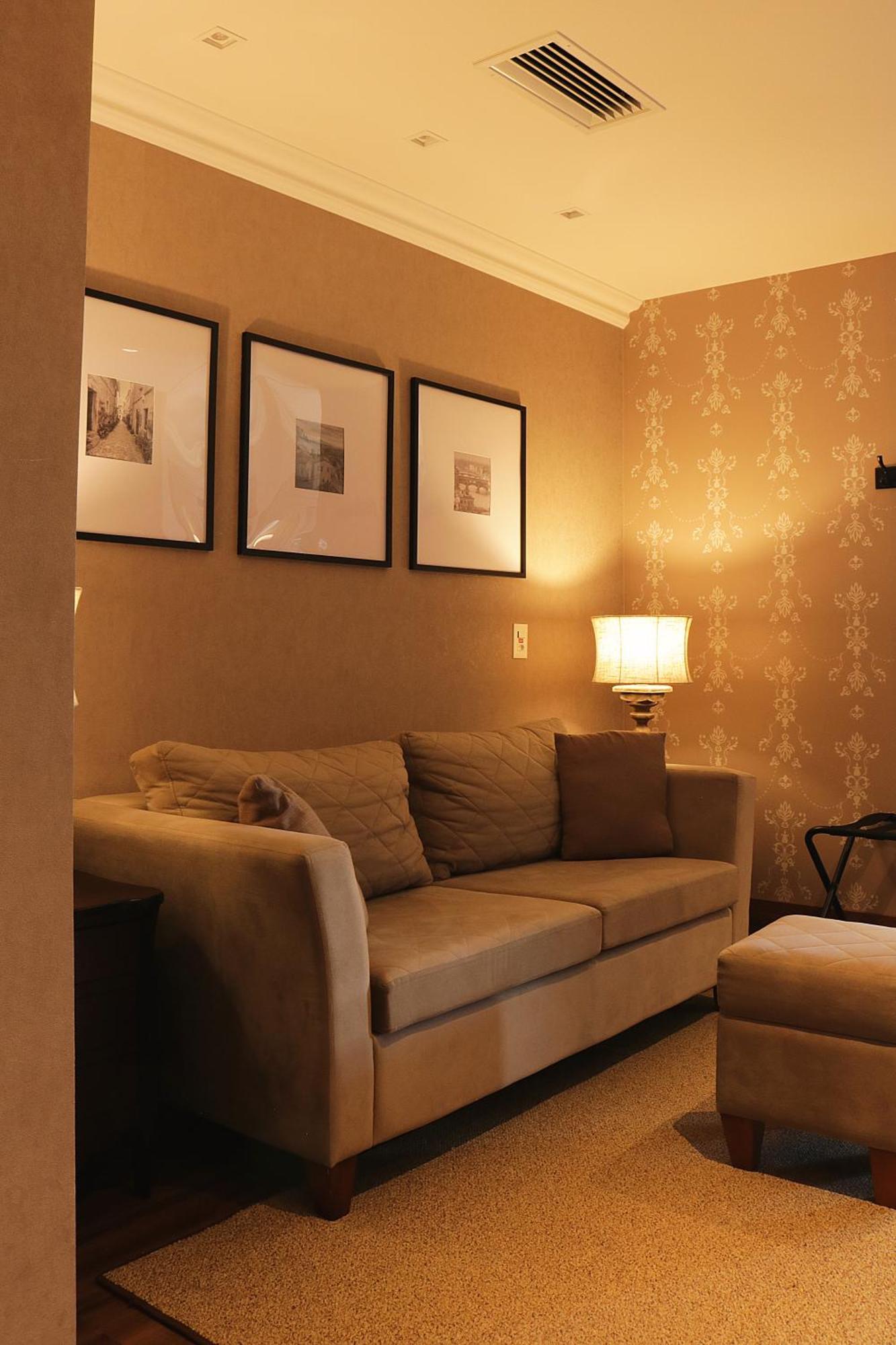 Buona Vitta Gramado Resort & Spa By Gramado Parks Zewnętrze zdjęcie The photo shows a cozy living room setting. There is a light-colored sofa adorned with several cushions, positioned against a wall that has a subtle pattern. Above the sofa, there are three framed artworks or photographs hanging on the wall. To the s
