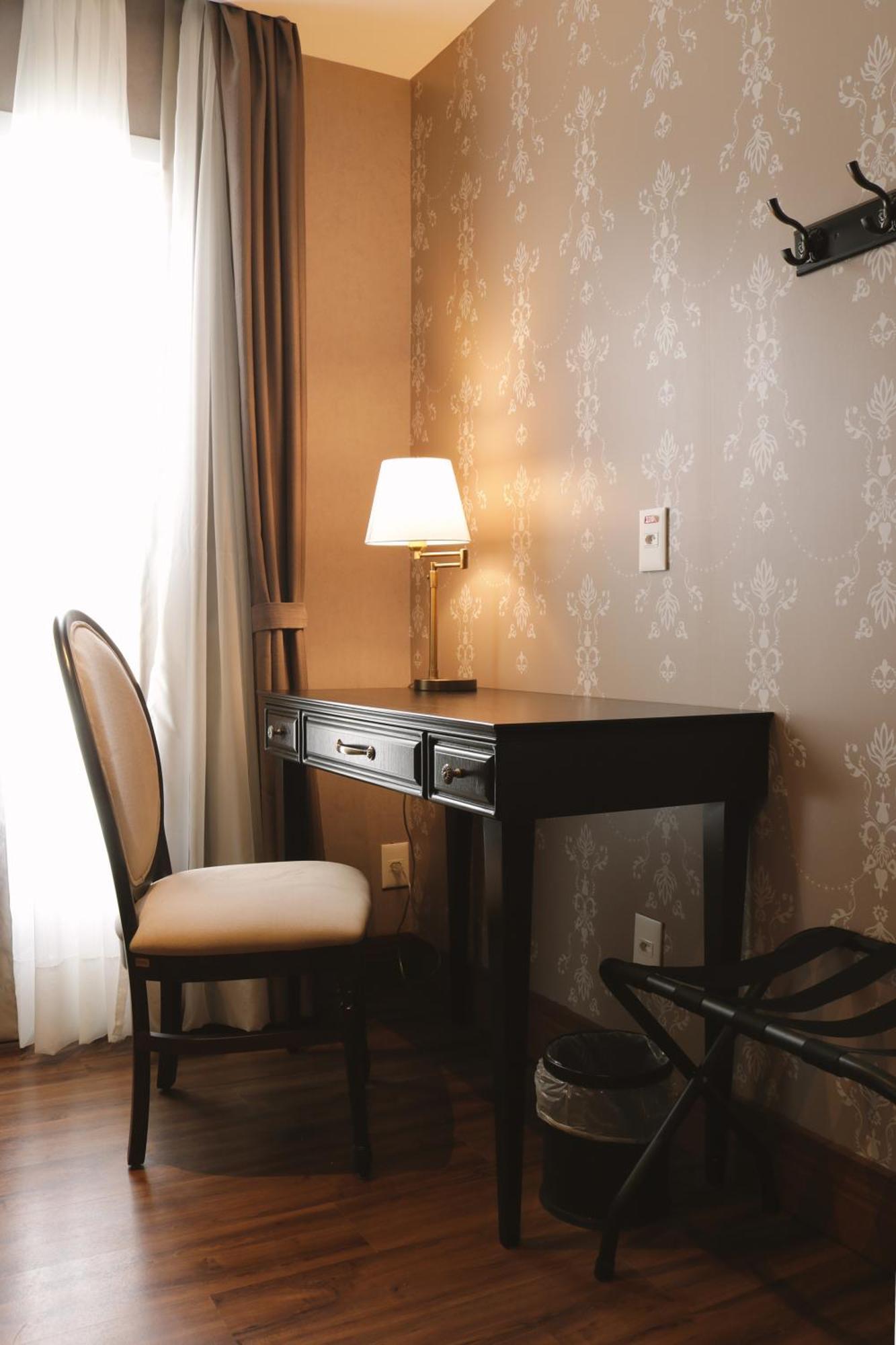 Buona Vitta Gramado Resort & Spa By Gramado Parks Zewnętrze zdjęcie The photo shows a small desk set against a wall with a decorative pattern. The desk is black with a few drawers and has a small lamp on it. Next to the desk, there is a chair with a light-colored cushion. Soft light is coming through a window, castin